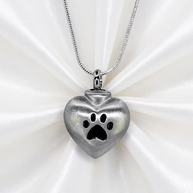 A silver heart-shaped pendant necklace with a paw print cutout, displayed on a white satin fabric background.