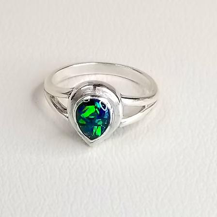 A sterling silver ring featuring a teardrop-shaped opal gemstone with vibrant green and blue play-of-color, set in a polished silver band on a textured off-white background.