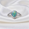 A silver ring featuring a teardrop-cut opal gemstone with vibrant iridescent colors, displayed on a draped white satin fabric background.