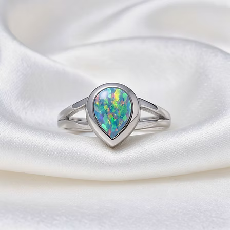 A silver ring featuring a teardrop-cut opal gemstone with vibrant iridescent colors, displayed on a draped white satin fabric background.