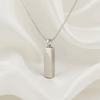 A silver-toned rectangular pendant necklace on a delicate chain, displayed against a draped white satin fabric background.