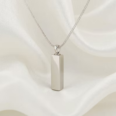 A silver-toned rectangular pendant necklace on a delicate chain, displayed against a draped white satin fabric background.
