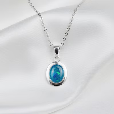 A silver oval pendant necklace featuring a vibrant blue iridescent opal stone, displayed on a draped white satin fabric background.