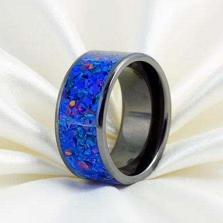 A polished dark gray metal ring with a vibrant blue iridescent inlay, resting on draped white satin fabric.