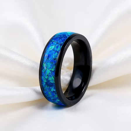 A black band ring featuring a vibrant blue and green iridescent inlay, displayed on a draped white satin fabric.
