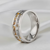 A polished silver-toned band ring with two inlaid textured stripes, one in warm gold and one in silver, resting on a soft white fabric background.