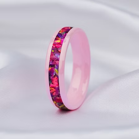 A pink band ring with a colorful floral inlay, resting on a soft white satin fabric background.
