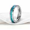 A polished titanium band ring with a vibrant blue and green inlay, resting on a draped white satin fabric against a white background.