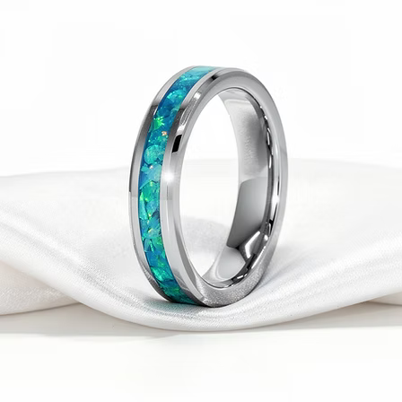 A polished titanium band ring with a vibrant blue and green inlay, resting on a draped white satin fabric against a white background.