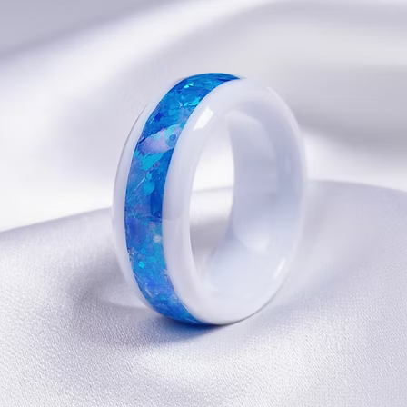 A white ceramic ring with a vibrant blue inlay, resting on a soft white fabric background.