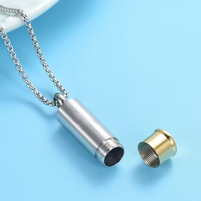 A silver cylindrical pendant necklace on a matching chain, shown with the threaded cap removed and placed beside it on a light blue background.