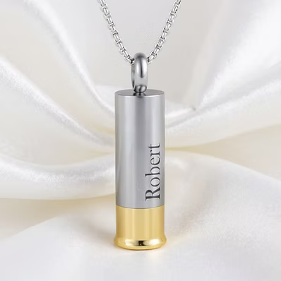 A silver cylindrical pendant necklace with a gold bottom, engraved with the name 'Robert', hanging from a silver chain against a draped white satin background.