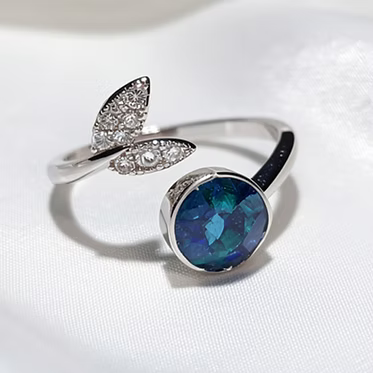 A silver open-band ring featuring two leaf-shaped settings with small clear stones and a large oval blue iridescent gemstone, photographed on a white fabric surface.