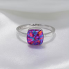 A silver band ring featuring a square cushion-cut opal gemstone with vibrant pink, purple, and blue iridescent play-of-color, displayed on a soft white satin fabric background.