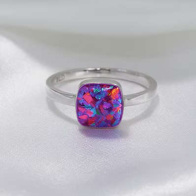 A silver band ring featuring a square cushion-cut opal gemstone with vibrant pink, purple, and blue iridescent play-of-color, displayed on a soft white satin fabric background.
