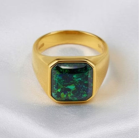 A gold signet ring featuring a square-cut green opal stone with vibrant blue and green flashes, displayed on a soft white satin fabric.