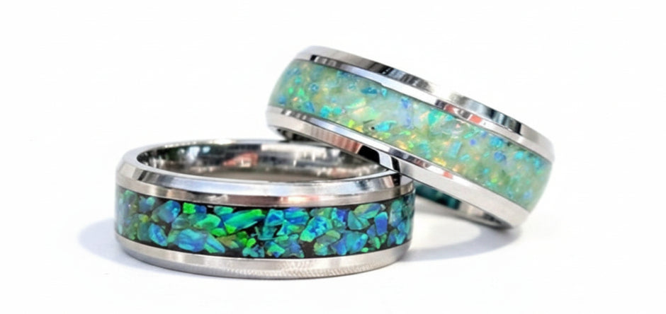 Two silver-toned wedding bands with vibrant green and blue opal inlays, resting on a draped white satin fabric.