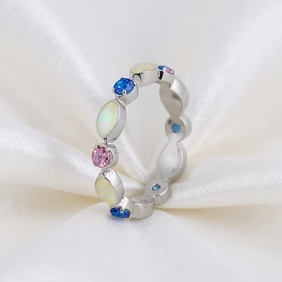 A silver band ring featuring a series of oval-cut iridescent opal-like stones and small blue and pink gemstones, displayed on a white satin fabric background.