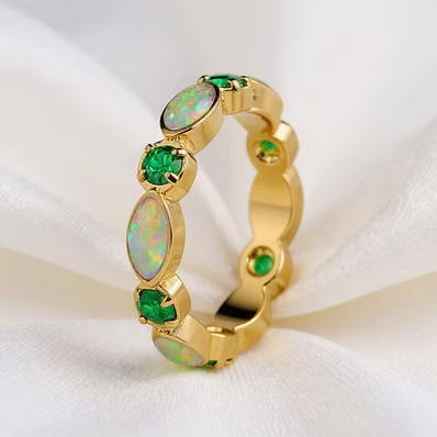 A gold ring featuring oval white opal and round green gemstone accents, displayed on a soft white fabric background.
