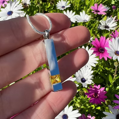A hand holding a silver-toned snake chain necklace with a rectangular pendant featuring a light blue center, a yellow crystal accent at the bottom, and a textured silver border, set against a background of green foliage and white and pink flowers.
