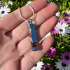 A silver-toned rectangular pendant necklace with a small red gemstone, held in a person's hand against a background of green foliage and white and pink flowers.