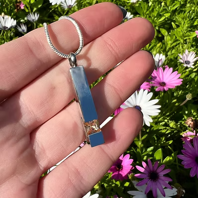 A rectangular blue pendant with a gold accent and a silver snake chain, held in a person's hand against a background of green foliage and pink and white flowers.