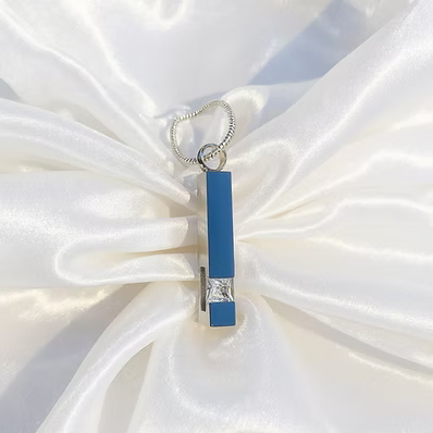 A silver-toned metal hair claw clip with a vertical blue enamel bar and a small clear rhinestone accent, displayed on a white satin fabric background.