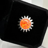A silver sunburst ring with a round orange opal center, displayed inside a black velvet jewelry box.