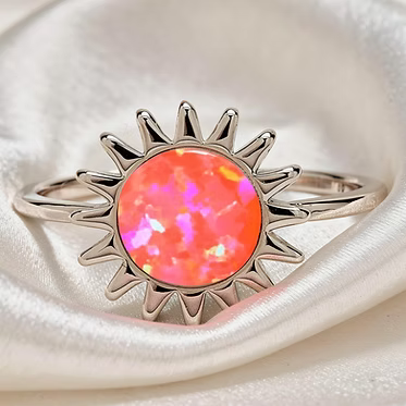 A silver sunburst ring featuring a vibrant, iridescent orange-pink opal gemstone, displayed on a soft white satin fabric.