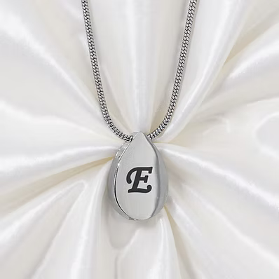 A silver teardrop-shaped pendant necklace with a black engraved letter 'E' on a silver chain, displayed on a white satin fabric background.