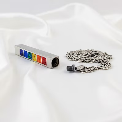 A silver metal cremation urn pendant with six colorful enamel inlays (blue, green, yellow, orange, red) and a matching silver chain, displayed on a draped white fabric background.
