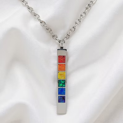 A silver-toned chakra necklace with a vertical rectangular pendant featuring seven colorful inlay squares in red, orange, yellow, green, blue, and purple, displayed on a white satin fabric background.