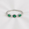 A silver ring featuring three marquise-cut green gemstones set in a wavy, flowing line, photographed on a soft white fabric background.