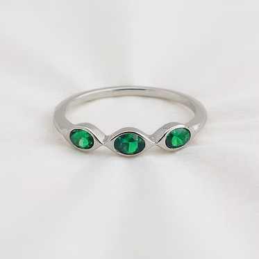 A silver ring featuring three marquise-cut green gemstones set in a wavy, flowing line, photographed on a soft white fabric background.