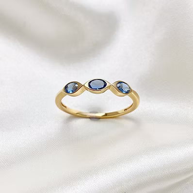 A gold ring with three oval blue gemstones, featuring a central larger stone and two smaller stones on either side, displayed on a draped white satin fabric.