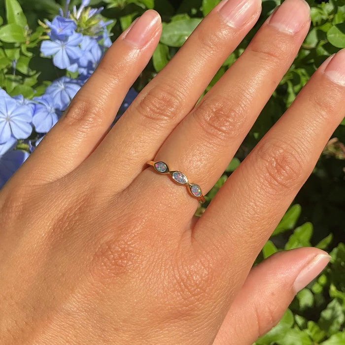 A close-up of a hand wearing a delicate three-stone ring outdoors, with green foliage and small blue flowers in the background.