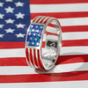 A silver-toned ring with a polished rectangular blue star inlay and red enamel stripe accents, photographed in front of an American flag background.