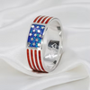 A silver band ring featuring an American flag design, with red inlaid stripes and a blue inlaid section with white star details, resting on a white satin fabric background.