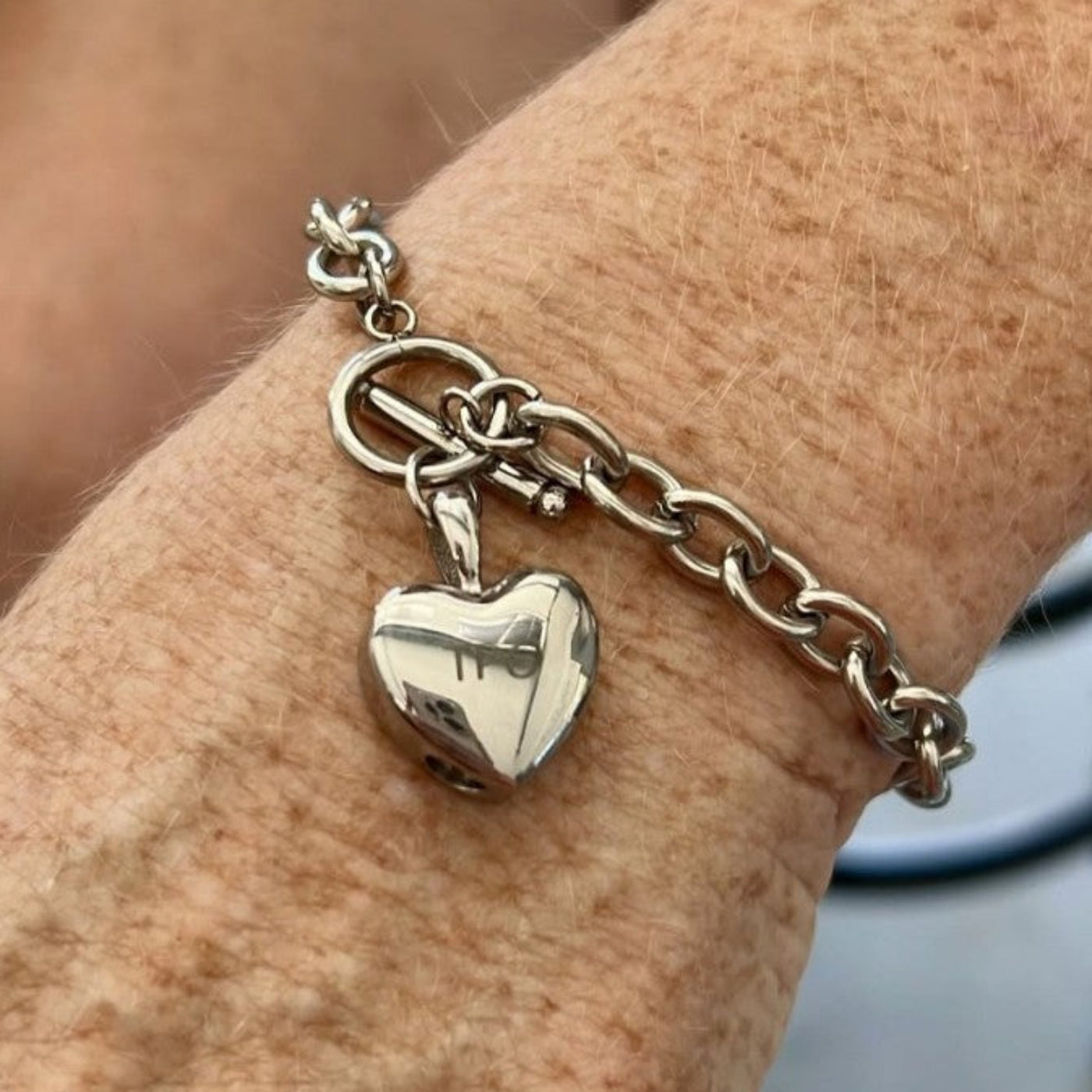 A silver charm bracelet with a heart-shaped pendant and a toggle clasp, shown worn on a person's wrist.