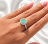A close-up of a hand wearing a silver halo ring with a cushion-cut green opal center stone, set against a soft white satin background.