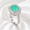 A silver ring featuring a cushion-cut green opal center stone surrounded by a halo of small clear stones, set on a white satin background.