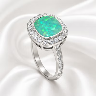 A silver ring featuring a cushion-cut green opal center stone surrounded by a halo of small clear stones, set on a white satin background.