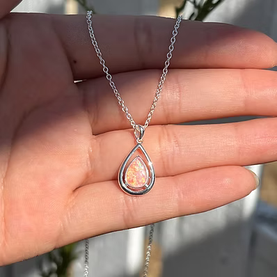 A silver teardrop pendant necklace with a pinkish-orange opal center, held in a person's hand.