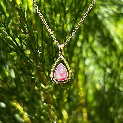 A gold teardrop pendant necklace with a pink stone, hanging against a blurred green foliage background.