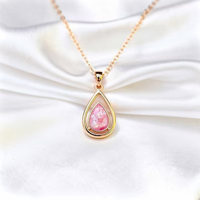 A gold teardrop pendant necklace with a pink iridescent stone, displayed on a draped white satin fabric background.