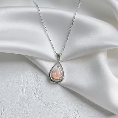 A silver teardrop pendant necklace with a milky white iridescent opal inlay, displayed on draped white satin fabric.