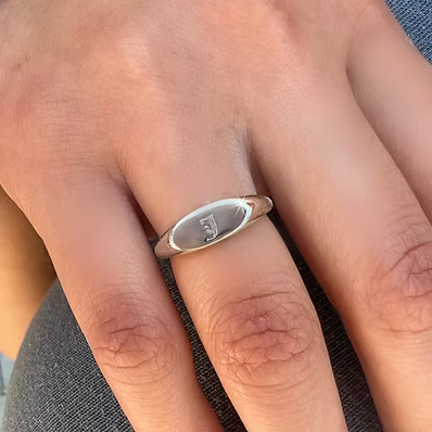 A close-up of a silver oval signet ring with a polished finish engraved with the initials 'J & E' on a person's ring finger.