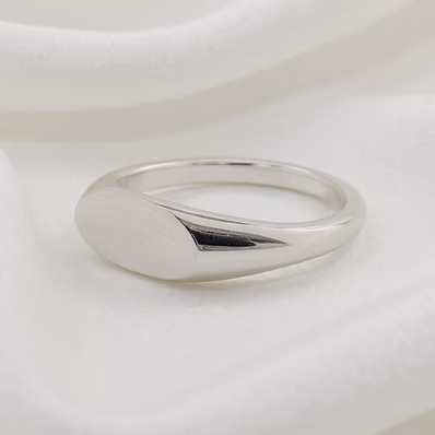A polished silver signet ring with a smooth, rounded oval face and a tapered band, resting on a soft, draped white fabric background.
