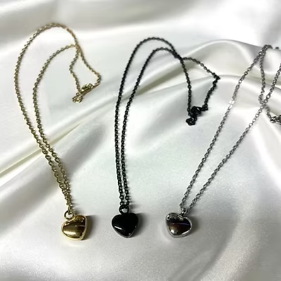 Three heart-shaped pendant necklaces displayed on draped white satin fabric, shown in gold, black, and silver tones.