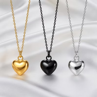 Three heart-shaped pendant necklaces displayed on draped satin fabric, featuring gold, black, and silver finishes.